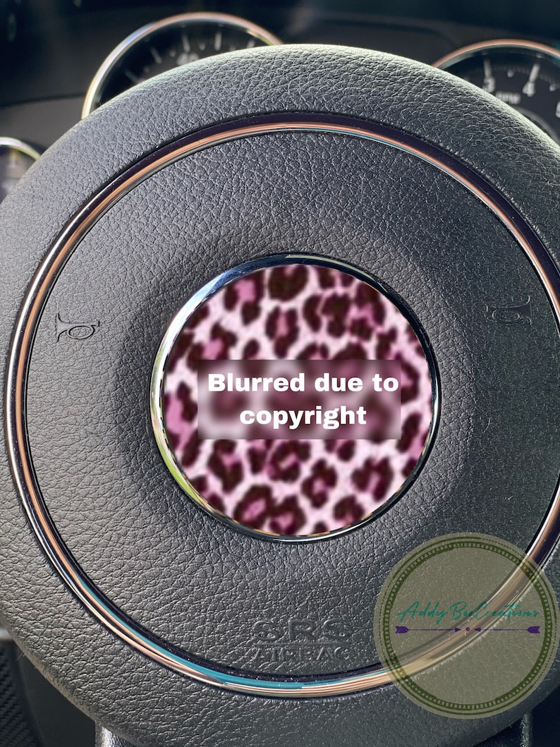 May include: A close-up of a black leather steering wheel with a pink and black leopard print design in the centre. The text "Blurred due to copyright" is visible in the centre of the design.