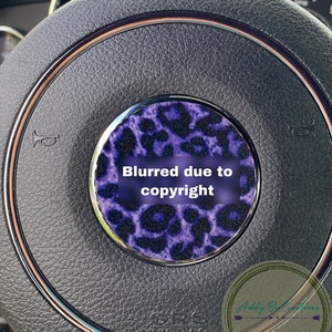 May include: A close-up of a black leather steering wheel with a purple and black leopard print design. The text "Blurred due to copyright" is visible on the design.