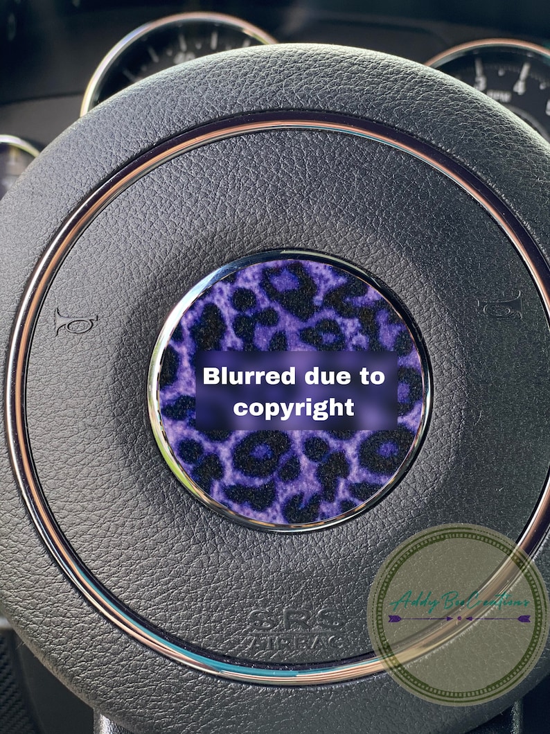 May include: A close-up of a black leather steering wheel with a purple and black leopard print design. The text "Blurred due to copyright" is visible on the design.