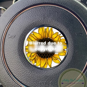 May include: A black leather steering wheel with a yellow sunflower design in the center. The sunflower has a honeycomb pattern in the center. The text "Blurred due to copyright" is visible on the sunflower.