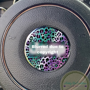 May include: A close-up of a black leather steering wheel with a decorative, round, teal, purple, and black leopard print sticker. The sticker has a white rectangle with the text "Blurred due to copyright".