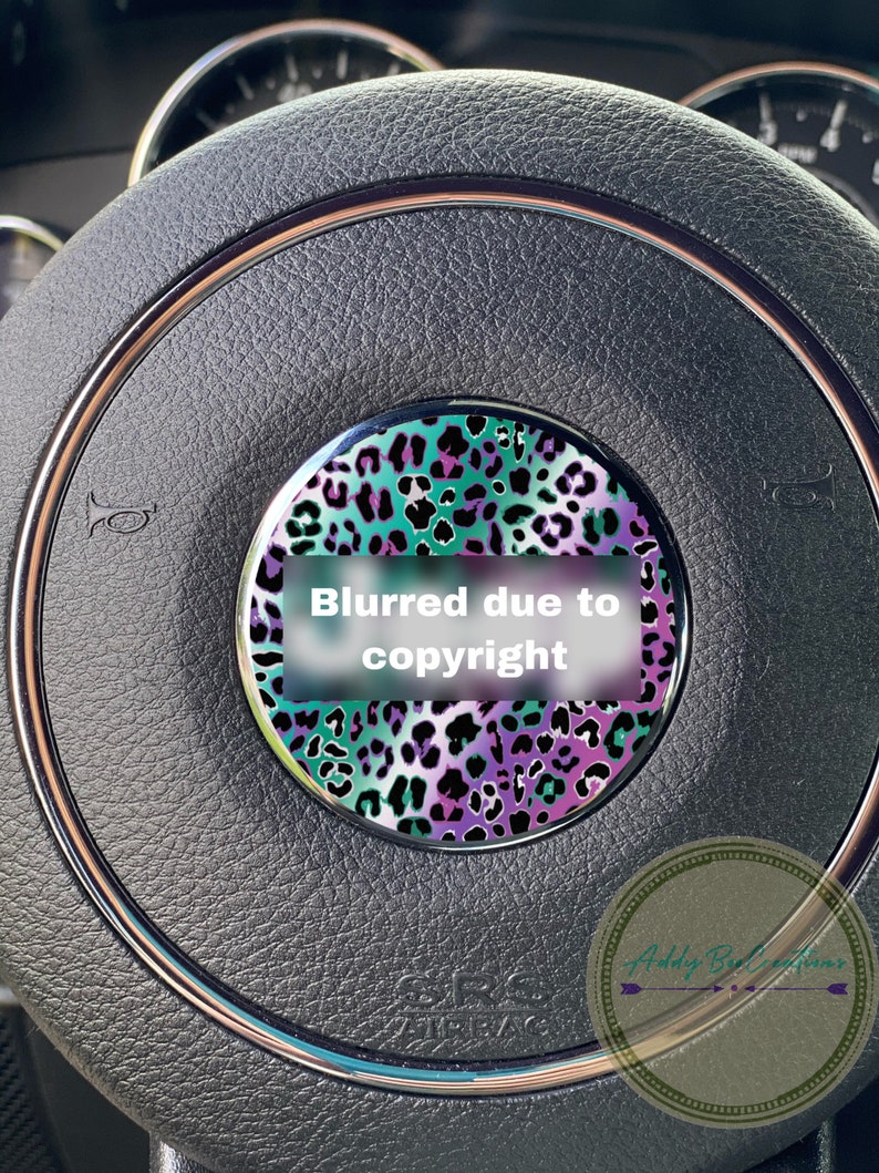 May include: A close-up of a black leather steering wheel with a decorative, round, teal, purple, and black leopard print sticker. The sticker has a white rectangle with the text "Blurred due to copyright".