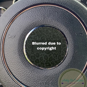 May include: Close-up of a black leather steering wheel with a rose gold trim. The centre of the steering wheel has a black and white leopard print design with the text "Blurred due to copyright".