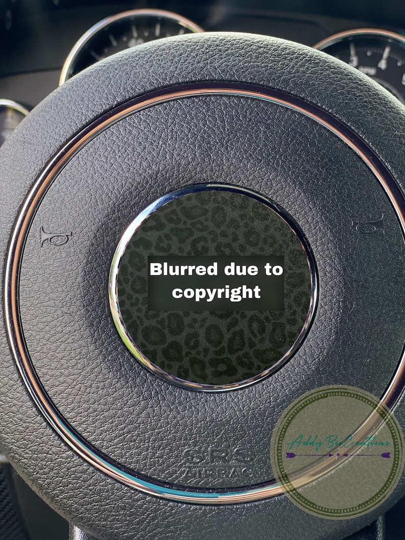 May include: Close-up of a black leather steering wheel with a rose gold trim. The centre of the steering wheel has a black and white leopard print design with the text "Blurred due to copyright".
