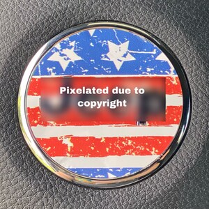 May include: A round, silver-rimmed sticker with a distressed American flag design. The sticker has red and white stripes and blue stars. The text "Pixelated due to copyright" is visible in the center of the sticker.