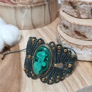 May include: A macrame bracelet featuring a central oval malachite stone with green banded patterns, set in a gold-toned metal frame. The band is crafted from dark gray cord with intricate knotting and small gold-toned beads integrated throughout.