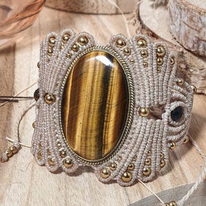 May include: A macrame bracelet featuring a large oval tiger's eye gemstone set in a brass-toned frame. The beige cord is intricately knotted and accented with small, round, metallic gold-coloured beads throughout the woven design.