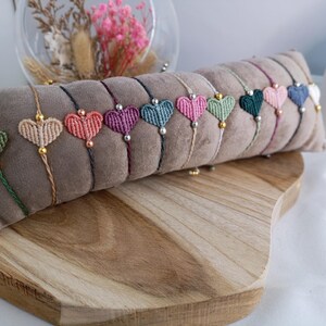 Peut inclure: Une collection de bracelets macramé en forme de cœur de différentes couleurs, dont vert, beige, rose, violet et bleu. Chaque bracelet est orné d'une perle dorée et est présenté sur un coussin en velours marron.