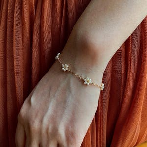 Peut inclure: Un bracelet d&eacute;licat en cha&icirc;ne dor&eacute;e avec de petites fleurs en perles blanches. Le bracelet est port&eacute; au poignet.