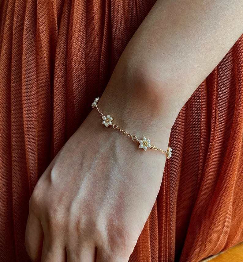Peut inclure: Un bracelet d&eacute;licat en cha&icirc;ne dor&eacute;e avec de petites fleurs en perles blanches. Le bracelet est port&eacute; au poignet.