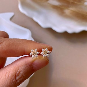 Peut inclure: Une paire de boucles d'oreilles en forme de fleur dor&eacute;es avec de petites perles blanches. Les boucles d'oreilles sont d&eacute;licates et f&eacute;minines.