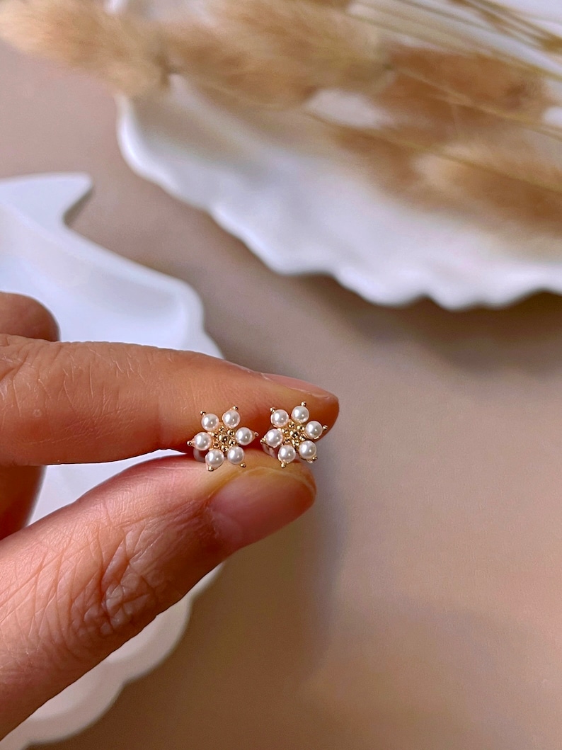 Peut inclure: Une paire de boucles d'oreilles en forme de fleur dor&eacute;es avec de petites perles blanches. Les boucles d'oreilles sont d&eacute;licates et f&eacute;minines.