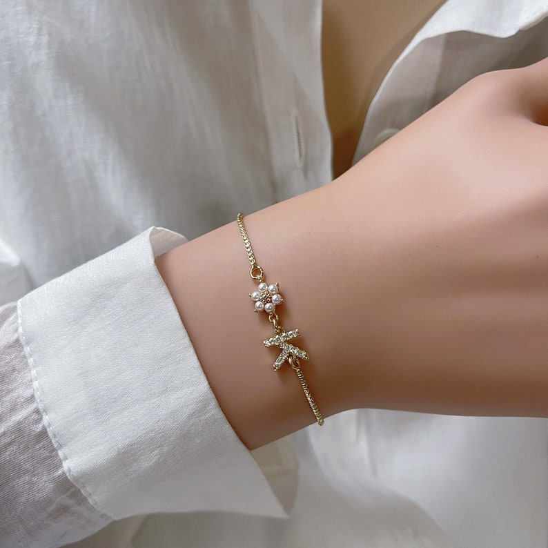 May include: A delicate gold chain bracelet with a small pearl flower charm and a letter 'K' charm. The bracelet is on a person's wrist.