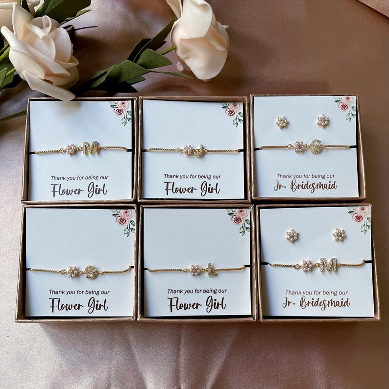 May include: Six small gift boxes with white lids and floral designs. Each box contains a gold chain bracelet with a charm and a message that says "Thank you for being our Flower Girl" or "Thank you for being our Jr. Bridesmaid".