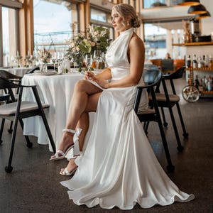 Könnte beinhalten: Eine Frau in einem weißen Halterneck-Kleid mit einem Oberschenkel-hohen Schlitz sitzt an einem Tisch in einem Restaurant. Sie trägt weiße Absätze und hat ihre Haare nach hinten gebunden. Der Tisch ist mit weißen Tischdecken und Blumen gedeckt.