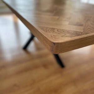 May include: Close-up of a wooden table top with a herringbone pattern. The table top is made of light brown wood and has a smooth, polished finish. The table top is supported by black metal legs.
