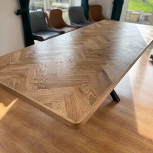 May include: A large wooden dining table with a herringbone pattern top. The table is made of light brown wood and has a black metal base.