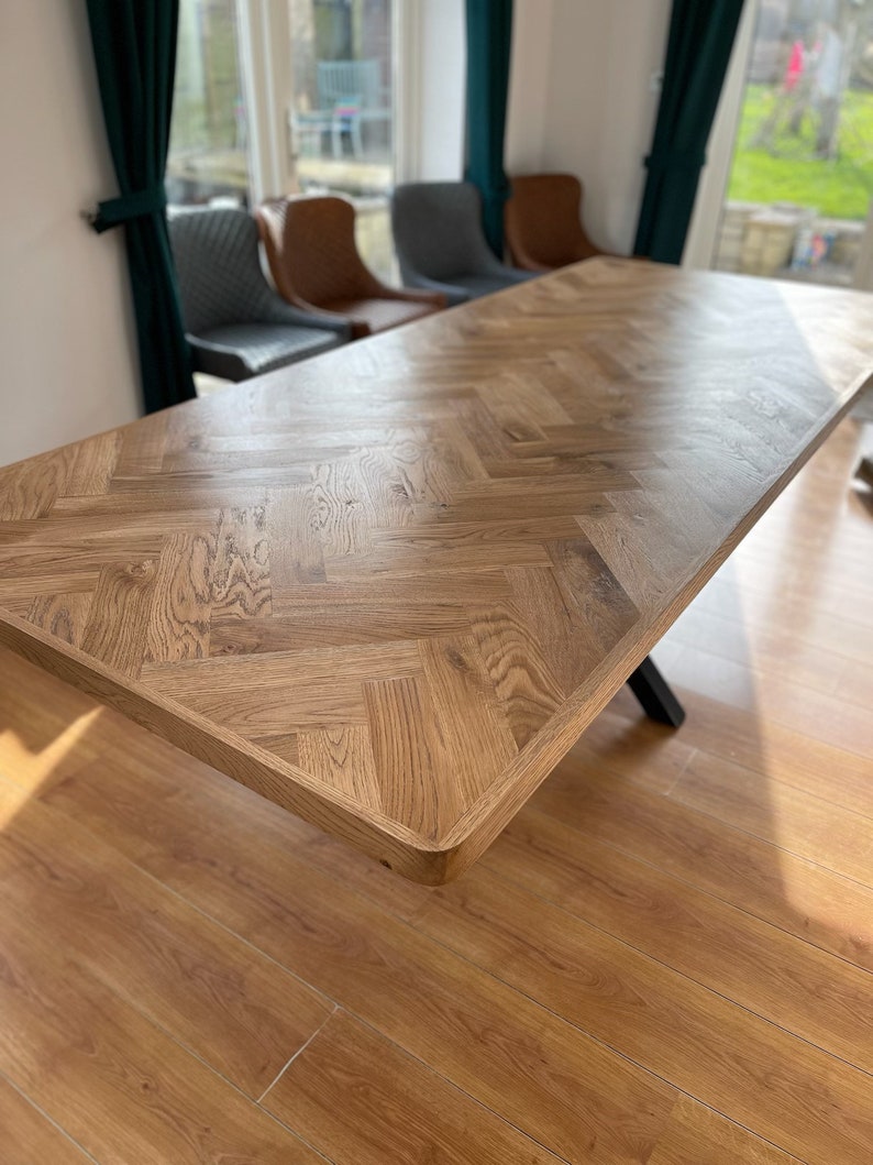 May include: A large wooden dining table with a herringbone pattern top. The table is made of light brown wood and has a black metal base.