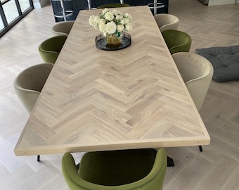 May include: A large wooden dining table with a herringbone pattern top. The table is surrounded by chairs with green and beige upholstery. A vase of white flowers sits in the center of the table.