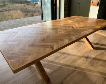 May include: A large wooden dining table with a herringbone pattern top and a simple X-shaped base. The table is made of natural wood with a light brown finish.