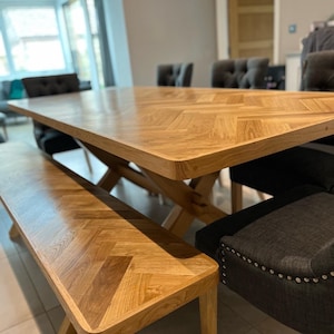 May include: A wooden dining table with a herringbone pattern top and a matching bench. The table is surrounded by chairs with dark grey upholstery and silver studs.