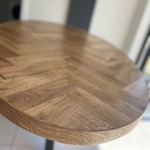 May include: Close-up of a round wooden table top with a herringbone pattern. The table top is a light brown color and has a smooth, polished finish.