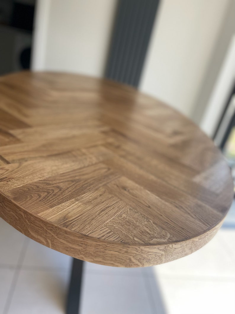 May include: Close-up of a round wooden table top with a herringbone pattern. The table top is a light brown color and has a smooth, polished finish.