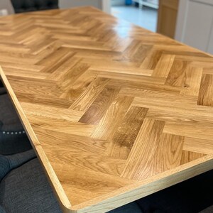 May include: A close-up of a wooden table top with a herringbone pattern. The table top is made of light brown wood and has a natural finish. The table top is surrounded by a dark grey fabric.