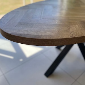 May include: Close-up of a wooden oval table top with a herringbone pattern. The table top is a light brown color and has a smooth, polished finish. The table top is supported by a black metal base.