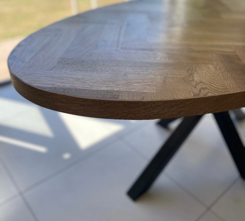 May include: Close-up of a wooden oval table top with a herringbone pattern. The table top is a light brown color and has a smooth, polished finish. The table top is supported by a black metal base.