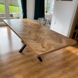May include: A large wooden dining table with a herringbone pattern top and black metal legs. The table is in a room with hardwood floors and a window in the background.