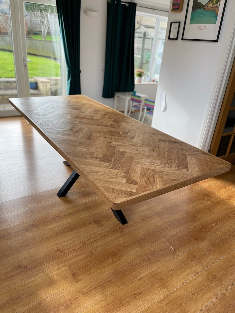 May include: A large wooden dining table with a herringbone pattern top and black metal legs. The table is in a room with hardwood floors and a window in the background.