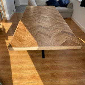 May include: A wooden table with a herringbone pattern top and a black metal base. The table is in a room with hardwood floors and a sofa in the background.