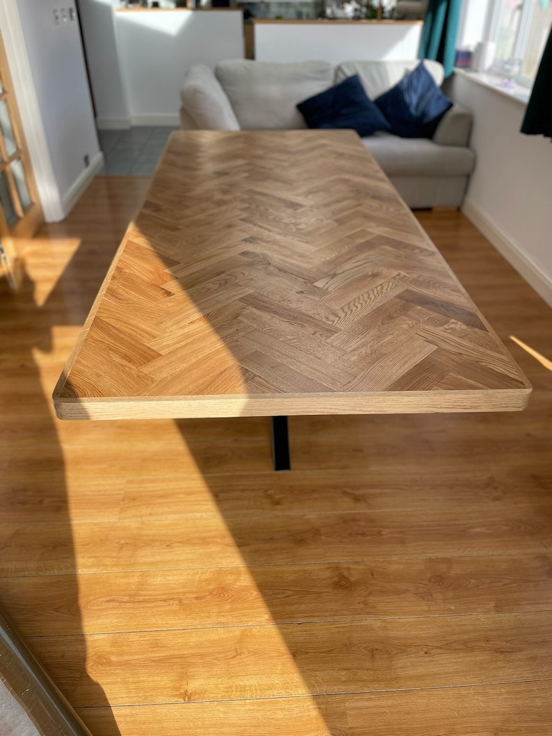 May include: A wooden table with a herringbone pattern top and a black metal base. The table is in a room with hardwood floors and a sofa in the background.