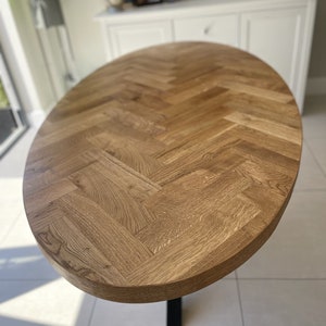 May include: A large oval wooden table top with a herringbone pattern. The table top is made of light brown wood and has a smooth, polished finish.