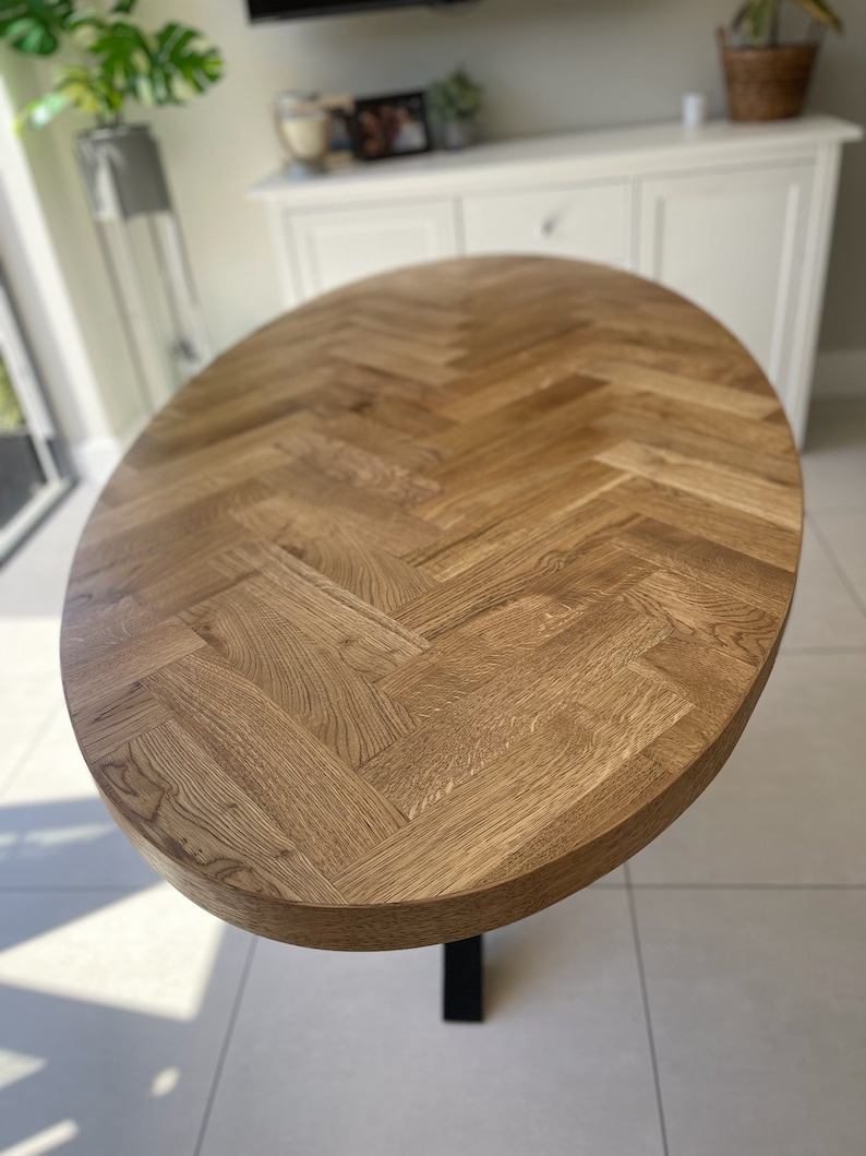 May include: A large oval wooden table top with a herringbone pattern. The table top is made of light brown wood and has a smooth, polished finish.