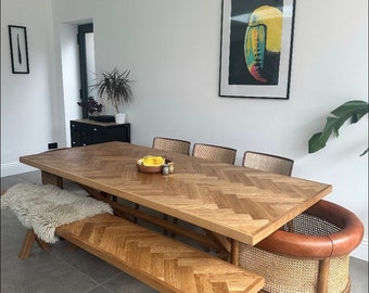 May include: A wooden dining table with a herringbone pattern top and a matching bench. The table is surrounded by chairs with woven seats and backs. A toucan artwork hangs on the wall behind the table.