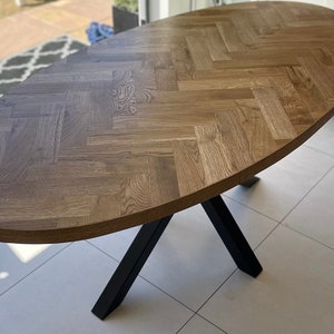 May include: A large oval wooden table with a herringbone pattern top and black metal legs. The table is on a light beige tiled floor.