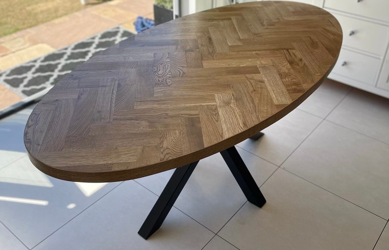 May include: A large oval wooden table with a herringbone pattern top and black metal legs. The table is on a light beige tiled floor.
