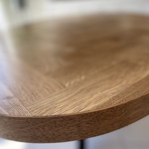 May include: Close-up of the edge of a round wooden table top with a natural wood grain finish. The table top is light brown in color and has a slightly rounded edge.
