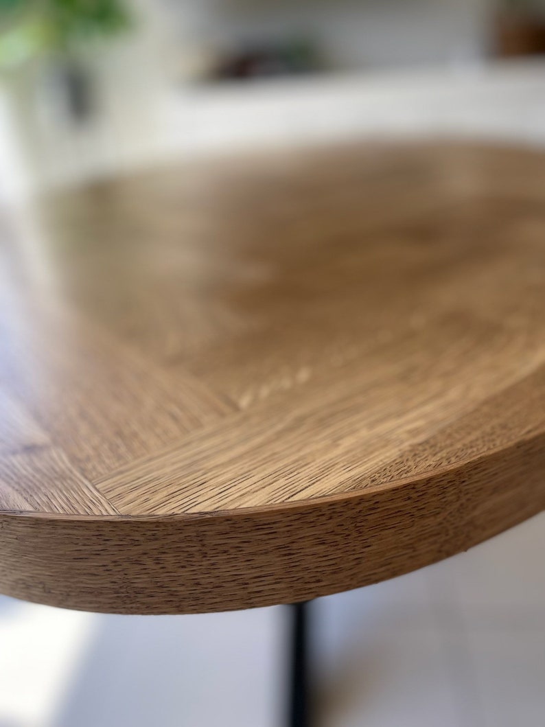 May include: Close-up of the edge of a round wooden table top with a natural wood grain finish. The table top is light brown in color and has a slightly rounded edge.