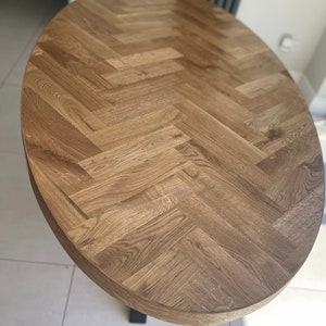 May include: Oval wooden table top with a herringbone pattern. The table top is made of light brown wood and has a smooth finish.