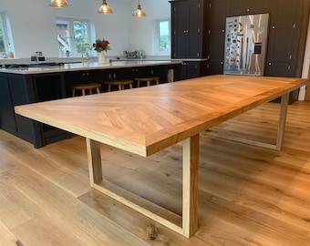 May include: A large wooden dining table with a chevron pattern top and a simple rectangular frame base. The table is made of light wood and has a natural finish. The table is in a modern kitchen with a white countertop and dark cabinets.