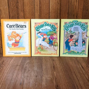 May include: Three children's books, two with yellow covers and one with an orange cover. The yellow covers feature illustrations of Cabbage Patch Kids characters. The orange cover features Care Bears characters. The titles of the books are "The Trouble With Timothy", "The Big Bicycle Race", and "Xavier's Fantastic Discovery".