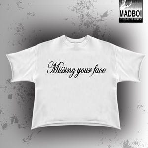 May include: White t-shirt featuring the cursive text "Missing your face". The top has a crew neck and short sleeves. A black and white logo reading "MADBOI PROJECT 0000" is in the upper right corner of the image.
