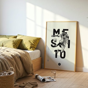 May include: A framed poster featuring a black and white image of a soccer player in action, with the name "MESSI" in large black letters. The poster is in a light-colored wooden frame and is displayed in a bedroom setting.