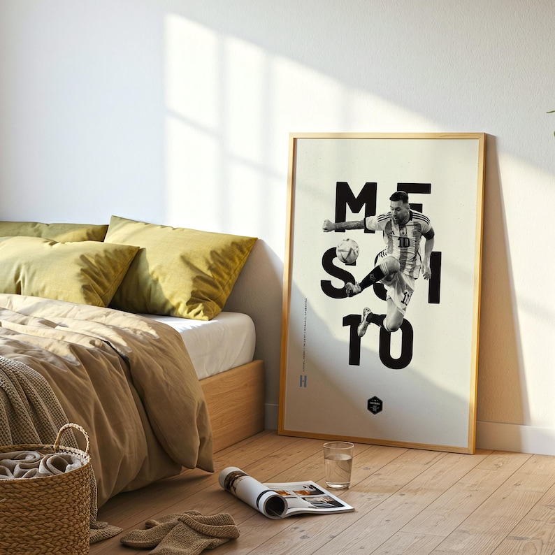 May include: A framed poster featuring a black and white image of a soccer player in action, with the name "MESSI" in large black letters. The poster is in a light-colored wooden frame and is displayed in a bedroom setting.