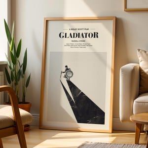 May include: Framed movie poster for the film "Gladiator" with a minimalist design. The poster features the film's title, credits, and a graphic of a gladiator. The poster is in a wooden frame and is displayed indoors.
