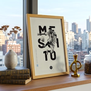 May include: Framed print featuring a black and white image of a soccer player kicking a ball, with the name "MESSI" and the number "10" in bold black letters. The frame is made of light-colored wood.