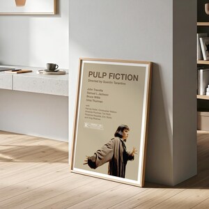 K&ouml;nnte beinhalten: Ein gerahmtes Filmplakat f&uuml;r "Pulp Fiction" mit einem Holzrahmen. Das Poster zeigt den Filmtitel, den Regisseur und die Besetzung, darunter John Travolta und Uma Thurman. Eine Frau in einem Mantel ist ebenfalls abgebildet.